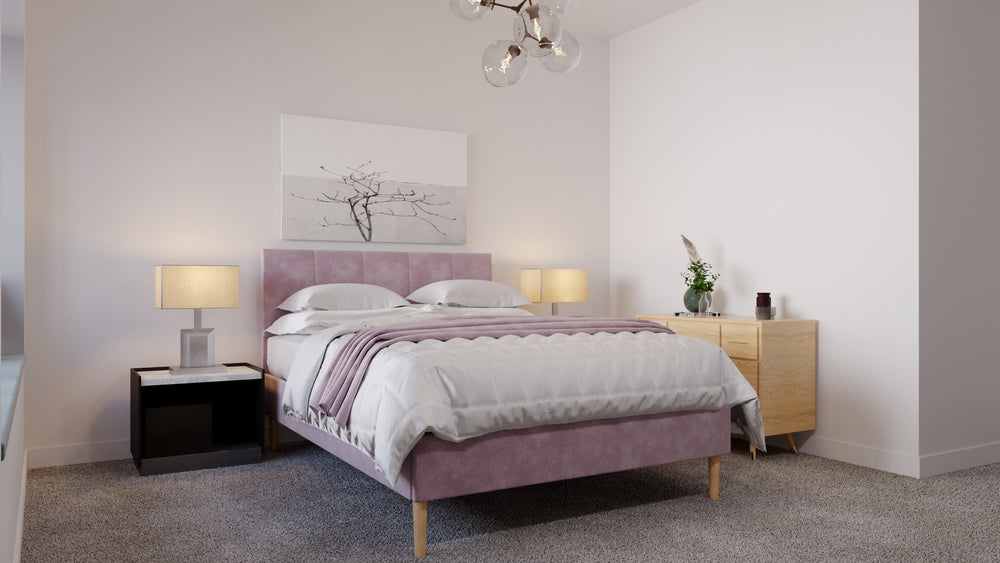 A pink fabric bed frame in the middle of a room which has grey carpet and white walls. The pillows and bedsheets are white. A throw hangs over the bed which is the same colour as the bed frame. 